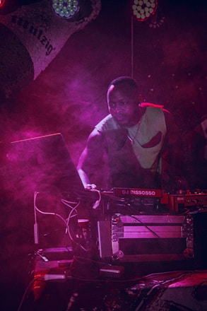 A DJ is focused on the music setup at a dimly lit event. Surrounded by a haze of pink and purple stage lights, he appears to be concentrating on the equipment, which includes a laptop and a DJ mixer. The atmosphere is vibrant and dynamic, with the lighting casting dramatic shadows.