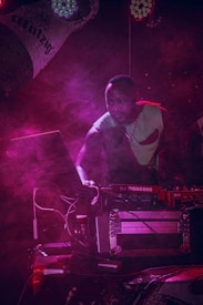 A DJ is focused on the music setup at a dimly lit event. Surrounded by a haze of pink and purple stage lights, he appears to be concentrating on the equipment, which includes a laptop and a DJ mixer. The atmosphere is vibrant and dynamic, with the lighting casting dramatic shadows.