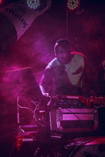 A DJ is focused on the music setup at a dimly lit event. Surrounded by a haze of pink and purple stage lights, he appears to be concentrating on the equipment, which includes a laptop and a DJ mixer. The atmosphere is vibrant and dynamic, with the lighting casting dramatic shadows.