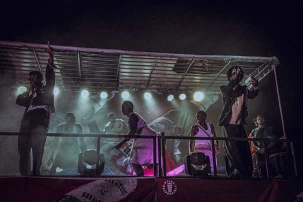 A group of performers is on a stage with bright lights illuminating the scene. The performers are actively singing or speaking into microphones, engaging with the audience energetically. The setting is dark, creating a contrast with the vibrant stage lights. The stage is partially covered with a roof structure, and there are several individuals wearing diverse outfits, suggesting a live performance or concert.
