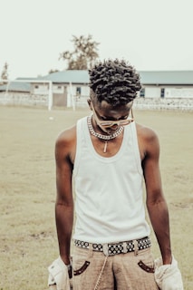 A young man wearing sunglasses and a white tank top stands outdoors. He has a necklace around his neck, a belt with metal details, and is holding a jacket. The background features grass and a blurry building.
