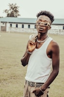 A fashionable individual stands outdoors, wearing sunglasses, a white tank top, and holding a jacket over their shoulder. They accessorize with a thick chain necklace and a studded belt. Their relaxed posture and confident expression suggest a sense of style and personality. The background features a grassy field and a building with multiple windows.