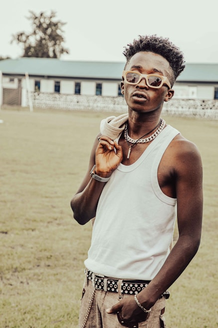A fashionable individual stands outdoors, wearing sunglasses, a white tank top, and holding a jacket over their shoulder. They accessorize with a thick chain necklace and a studded belt. Their relaxed posture and confident expression suggest a sense of style and personality. The background features a grassy field and a building with multiple windows.