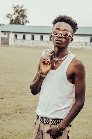 A fashionable individual stands outdoors, wearing sunglasses, a white tank top, and holding a jacket over their shoulder. They accessorize with a thick chain necklace and a studded belt. Their relaxed posture and confident expression suggest a sense of style and personality. The background features a grassy field and a building with multiple windows.