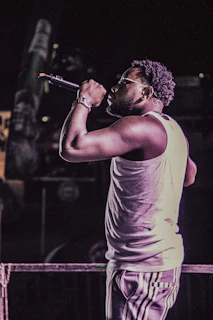A performer, wearing a sleeveless shirt and striped pants, holds a microphone while singing or speaking on stage. The scene is dimly lit, creating a focus on the subject with a subtle spotlight effect.