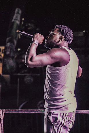 A performer, wearing a sleeveless shirt and striped pants, holds a microphone while singing or speaking on stage. The scene is dimly lit, creating a focus on the subject with a subtle spotlight effect.
