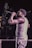 A performer, wearing a sleeveless shirt and striped pants, holds a microphone while singing or speaking on stage. The scene is dimly lit, creating a focus on the subject with a subtle spotlight effect.