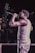 A performer, wearing a sleeveless shirt and striped pants, holds a microphone while singing or speaking on stage. The scene is dimly lit, creating a focus on the subject with a subtle spotlight effect.