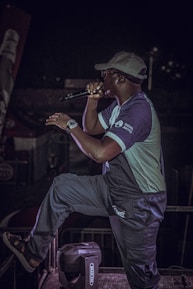 A person wearing a cap and glasses is performing on stage with a microphone. They are dressed casually in a two-toned outfit, and there is stage equipment visible in the background. The person appears to be in the middle of a dynamic performance, possibly singing or speaking.