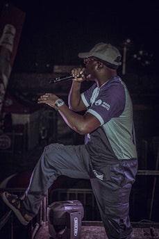A person wearing a cap and glasses is performing on stage with a microphone. They are dressed casually in a two-toned outfit, and there is stage equipment visible in the background. The person appears to be in the middle of a dynamic performance, possibly singing or speaking.