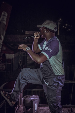 A person wearing a cap and glasses is performing on stage with a microphone. They are dressed casually in a two-toned outfit, and there is stage equipment visible in the background. The person appears to be in the middle of a dynamic performance, possibly singing or speaking.