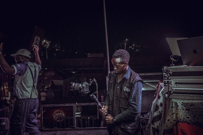 A person is holding a professional camera on a stabilizer, positioned near audio-visual equipment, with another person raising their arms nearby. The background suggests a lively event or performance setting, possibly a concert.