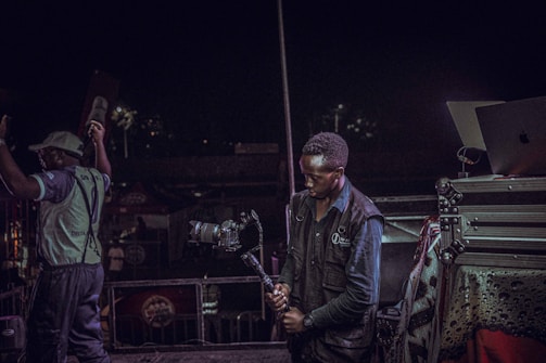 A person is holding a professional camera on a stabilizer, positioned near audio-visual equipment, with another person raising their arms nearby. The background suggests a lively event or performance setting, possibly a concert.