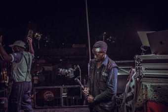 A person is holding a professional camera on a stabilizer, positioned near audio-visual equipment, with another person raising their arms nearby. The background suggests a lively event or performance setting, possibly a concert.