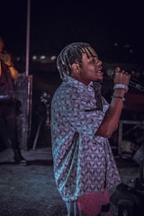 A person with braided hair is passionately singing into a microphone on a dimly lit stage. They are wearing a patterned shirt and bracelets, and the background features another person and stage equipment.