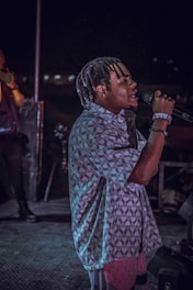 A person with braided hair is passionately singing into a microphone on a dimly lit stage. They are wearing a patterned shirt and bracelets, and the background features another person and stage equipment.