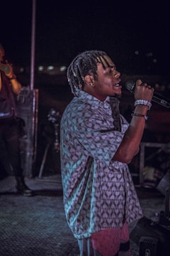 A person with braided hair is passionately singing into a microphone on a dimly lit stage. They are wearing a patterned shirt and bracelets, and the background features another person and stage equipment.