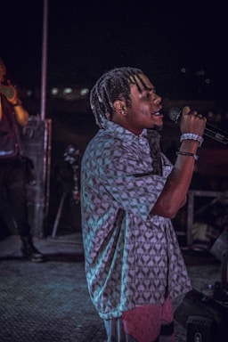 A person with braided hair is passionately singing into a microphone on a dimly lit stage. They are wearing a patterned shirt and bracelets, and the background features another person and stage equipment.