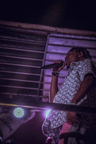 A person with a microphone in hand appears to be singing or speaking, standing on a stage-like area. The setting has a dim, moody lighting with purple hues and there are bright stage lights in the background, casting a glow.