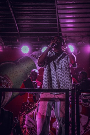 A performer on stage is holding a microphone, with bright stage lights behind creating a silhouette effect. The setting has a colorful backdrop with patterns and designs. People in the background appear to be part of a band or crew.