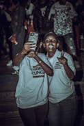 Two people are taking a selfie at an event. They are both wearing matching light-colored shirts with text on them. The background is dark with other people visible, suggesting a lively atmosphere.