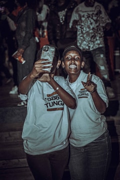Two people are taking a selfie at an event. They are both wearing matching light-colored shirts with text on them. The background is dark with other people visible, suggesting a lively atmosphere.