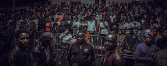 A large crowd gathers indoors, with a group of people in the foreground. Many are dressed casually, and some hold drinks or take photos. The lighting is dim, suggesting an evening event or party atmosphere.