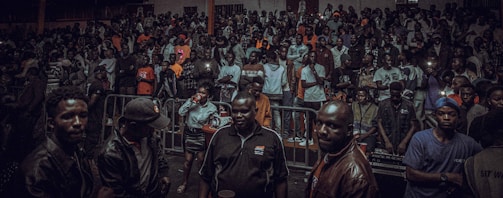 A large crowd gathers indoors, with a group of people in the foreground. Many are dressed casually, and some hold drinks or take photos. The lighting is dim, suggesting an evening event or party atmosphere.