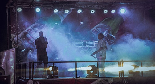 A dimly lit stage features performers surrounded by colorful fog, with bright stage lights overhead. The backdrop displays large images of beverage bottles, creating a lively and energetic atmosphere.