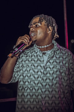 A person holding a microphone is seen performing. The individual has braided hair and is wearing a patterned shirt with heart and spade designs. They have a necklace with white beads and are captured in a dynamic pose.