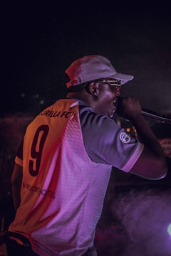A performer wearing a sports jersey with the number 9 and a cap is energetically singing into a microphone. The scene is set in a dimly lit environment with a mix of colorful lights creating a vibrant atmosphere.