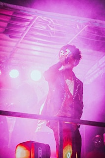 A person with curly hair is on a stage, holding a microphone, surrounded by vibrant pink and purple lighting. Smoke or fog adds to the atmosphere, and the person is wearing a stylish jacket with chains and a patterned shirt. Bright lights in the background enhance the stage presence.