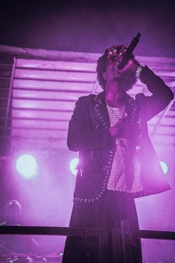 A performer is passionately singing into a microphone on stage. The atmosphere is enhanced by vibrant stage lights creating a moody and dramatic ambiance. The performer is wearing a stylish outfit with metal studs and a mask or adornment on their face.