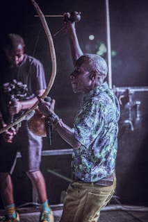 A person wearing a patterned shirt and holding a traditional musical instrument appears to be performing or playing. Another person stands nearby holding equipment that might be a camera. The setting is a dimly lit stage or performance area, suggesting a live music event.