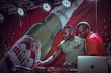 Two men are standing in front of a large promotional banner featuring a beer bottle. They are interacting with DJ equipment and a laptop while under spotlights. One man appears to be demonstrating something to the other.