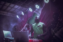 A DJ stands behind a laptop and a DJ controller with red branding, under colorful stage lighting. The background features a large, artistic graphic and smoke rises around the scene, suggesting an energetic atmosphere.