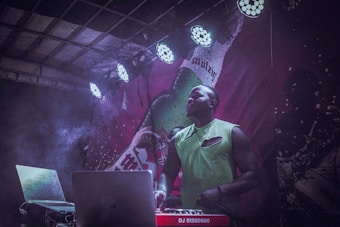 A DJ stands behind a laptop and a DJ controller with red branding, under colorful stage lighting. The background features a large, artistic graphic and smoke rises around the scene, suggesting an energetic atmosphere.