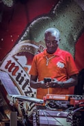 A person in a red polo shirt looks at a phone while standing in front of a large beer bottle advertisement. DJ equipment is set up in the foreground, and there is a logo or name tag on the shirt.