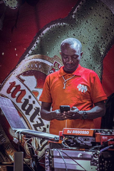A person in a red polo shirt looks at a phone while standing in front of a large beer bottle advertisement. DJ equipment is set up in the foreground, and there is a logo or name tag on the shirt.