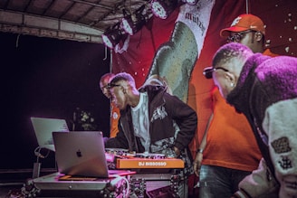 A group of people is gathered around a DJ setup, with one person actively operating the turntables. The scene includes two laptops, and some stage lighting is visible overhead. The backdrop features a large image of a beverage bottle.