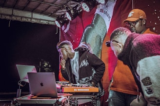 Three men stand around a DJ setup on stage. The person on the left is using a laptop, while another person appears to be overseeing the equipment. They are surrounded by a vivid, colorful backdrop with a bright red banner featuring an image of a beer bottle. The setting suggests a lively, nighttime event.