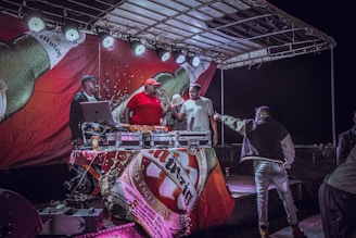 A group of people is gathered on a stage with DJ equipment. The stage has a large banner featuring a beer bottle and lights above. One person in a red shirt and cap operates the DJ equipment, while others are engaged in conversation or handing items to each other.