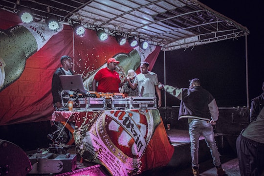 A group of people is gathered on a stage with DJ equipment. The stage has a large banner featuring a beer bottle and lights above. One person in a red shirt and cap operates the DJ equipment, while others are engaged in conversation or handing items to each other.