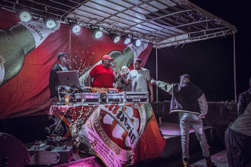 A group of people is gathered on a stage with DJ equipment. The stage has a large banner featuring a beer bottle and lights above. One person in a red shirt and cap operates the DJ equipment, while others are engaged in conversation or handing items to each other.