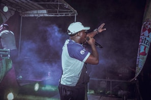 A performer is on stage holding a microphone, gesturing towards the audience. The scene is set under a stage canopy, with colorful lights and fog adding a dramatic effect. A promotional banner is visible on the side.