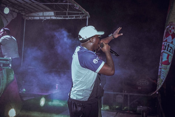 A performer is on stage holding a microphone, gesturing towards the audience. The scene is set under a stage canopy, with colorful lights and fog adding a dramatic effect. A promotional banner is visible on the side.