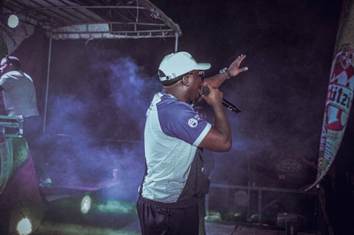 A performer is on stage holding a microphone, gesturing towards the audience. The scene is set under a stage canopy, with colorful lights and fog adding a dramatic effect. A promotional banner is visible on the side.