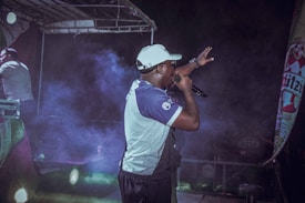 A performer is on stage holding a microphone, gesturing towards the audience. The scene is set under a stage canopy, with colorful lights and fog adding a dramatic effect. A promotional banner is visible on the side.