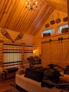 A cozy wooden cabin interior with high wooden ceilings and walls. There is a bed in the foreground with luggage and clothes on it. On the left, there are long striped curtains covering large windows. Snowshoes are mounted on the wall above the windows. A wooden chair and various items are placed beside the windows. The back wall features two small, high windows with baskets on top. There is also a set of barn-style sliding doors in the back.