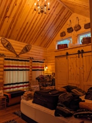 A cozy wooden cabin interior with high wooden ceilings and walls. There is a bed in the foreground with luggage and clothes on it. On the left, there are long striped curtains covering large windows. Snowshoes are mounted on the wall above the windows. A wooden chair and various items are placed beside the windows. The back wall features two small, high windows with baskets on top. There is also a set of barn-style sliding doors in the back.